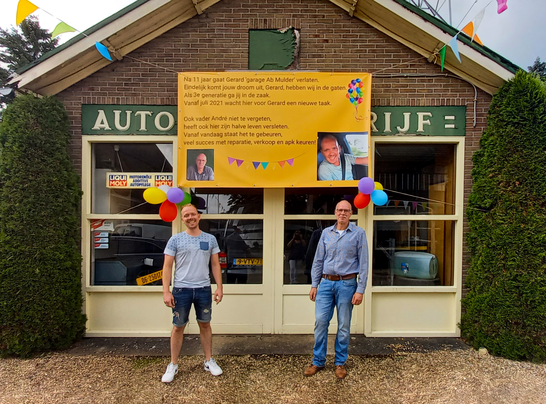 Twee mannen voor de oude gevel van de garage, met boven de ramen een geel spandoek van de derde generatie en de groene Autobedrijf-plaat