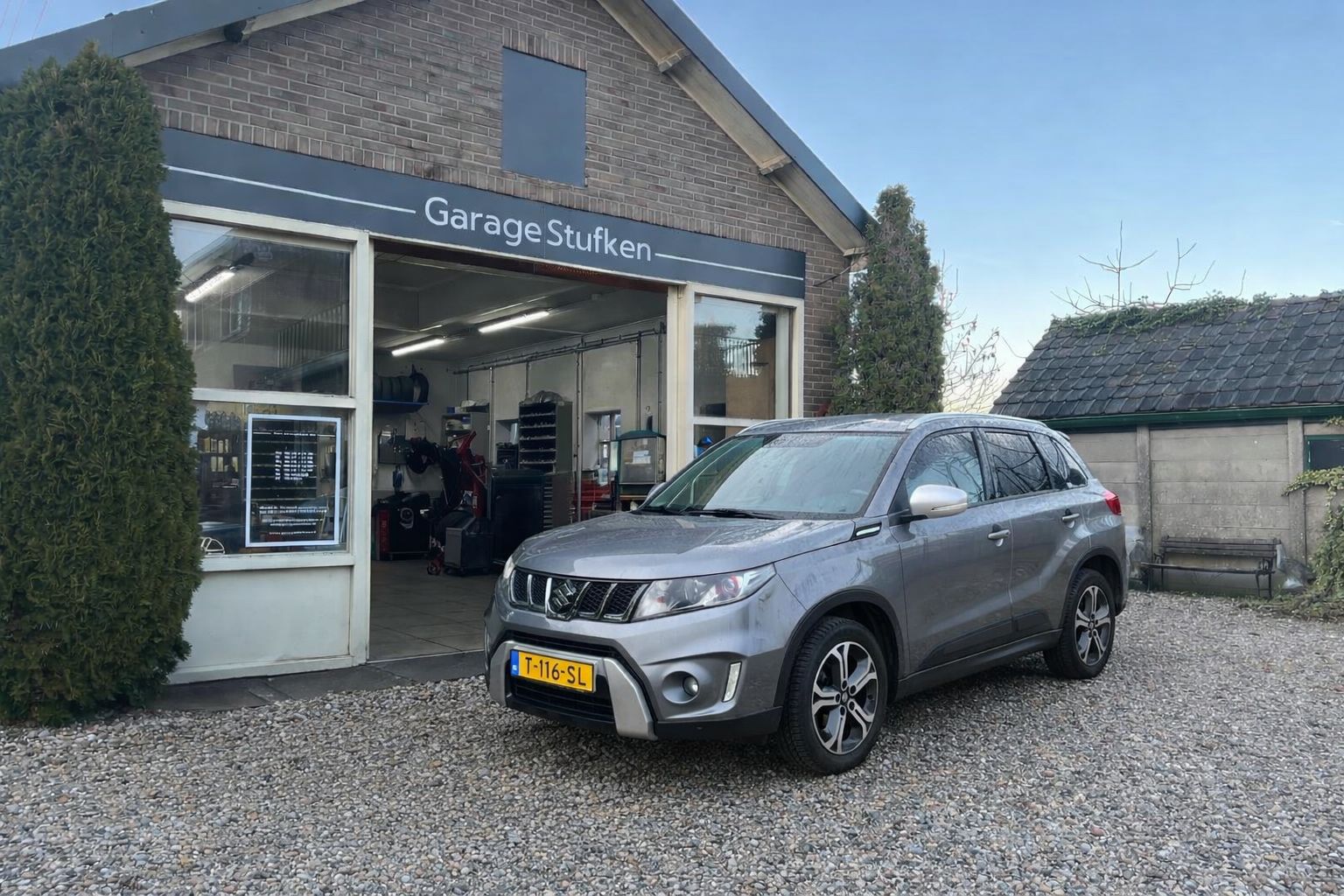 Grijze Suzuki Vitara voor de gevel van Garage Stufken, met de open werkplaats erachter